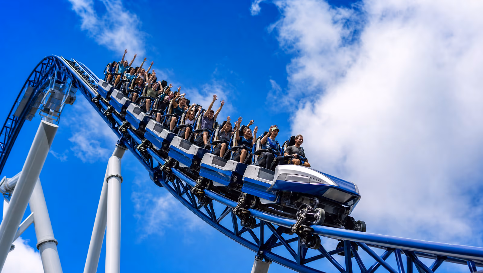 Riders with arms raised on a blue roller coaster against a blue sky